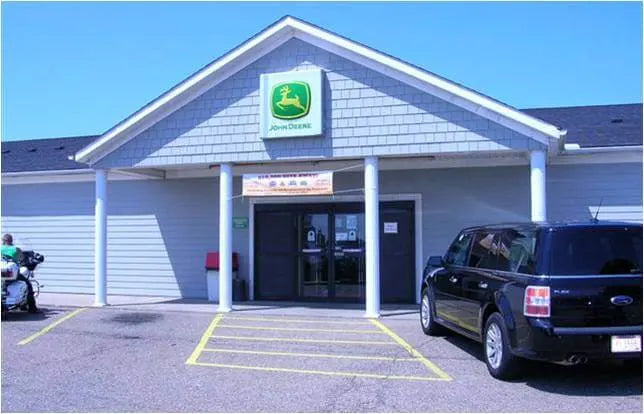 Single-story gray building with white columns supporting a triangular roof displaying a green and yellow John Deere logo above double glass doors, with a black SUV parked in front under clear blue sky.