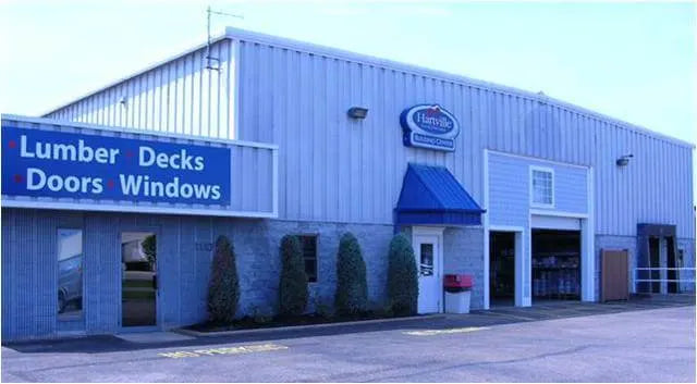Exterior view of a light gray metal building with a blue awning entrance and signage advertising lumber, decks, doors, and windows, set against a clear sky.