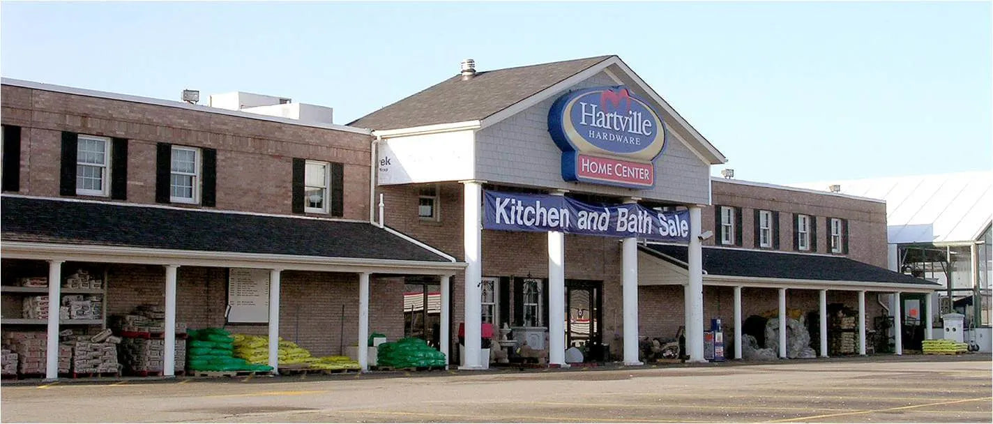 Wide front view of a two-story brick building with white trim and a Hartville Hardware sign above the entrance, featuring a banner for a kitchen and bath sale and stacks of building supplies along the covered walkway.