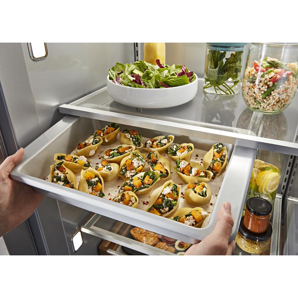 Hands sliding a bakeware tray with stuffed shells into under-shelf prep zone of built-in refrigerator interior
