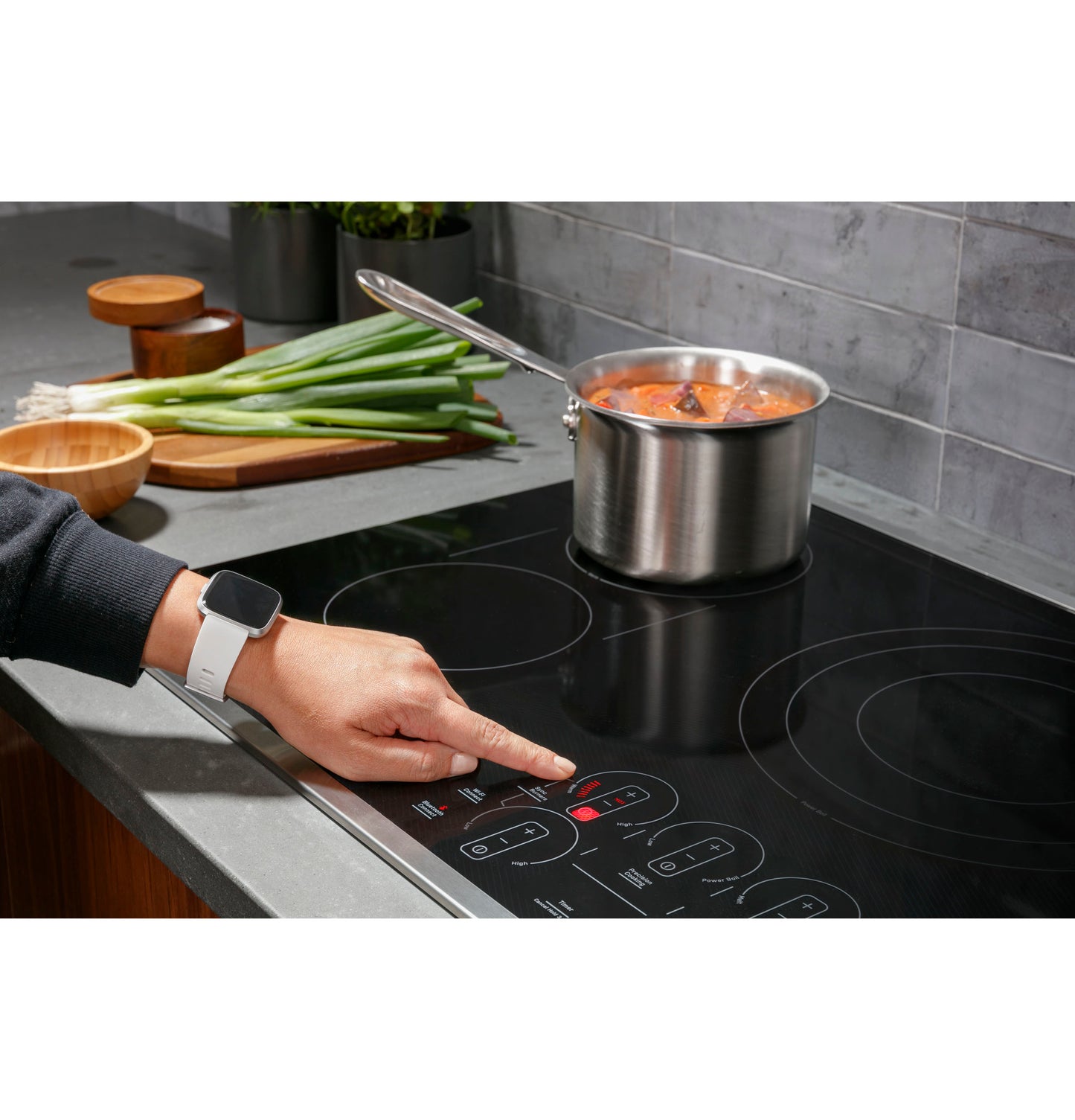 Hand adjusting touch control on black electric cooktop with stainless steel pot cooking tomato sauce nearby in modern kitchen