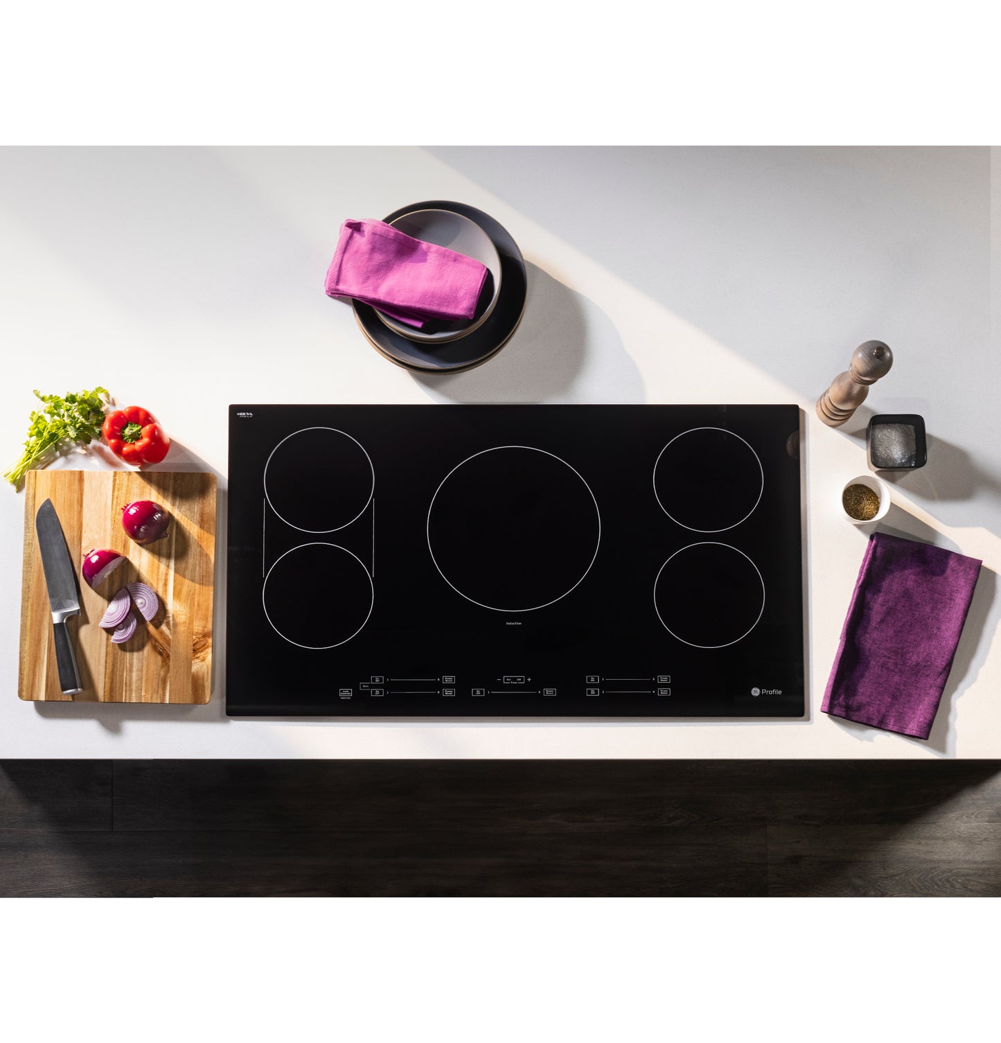Sleek black touch control induction cooktop with five cooking zones on white countertop with fresh vegetables and kitchen tools