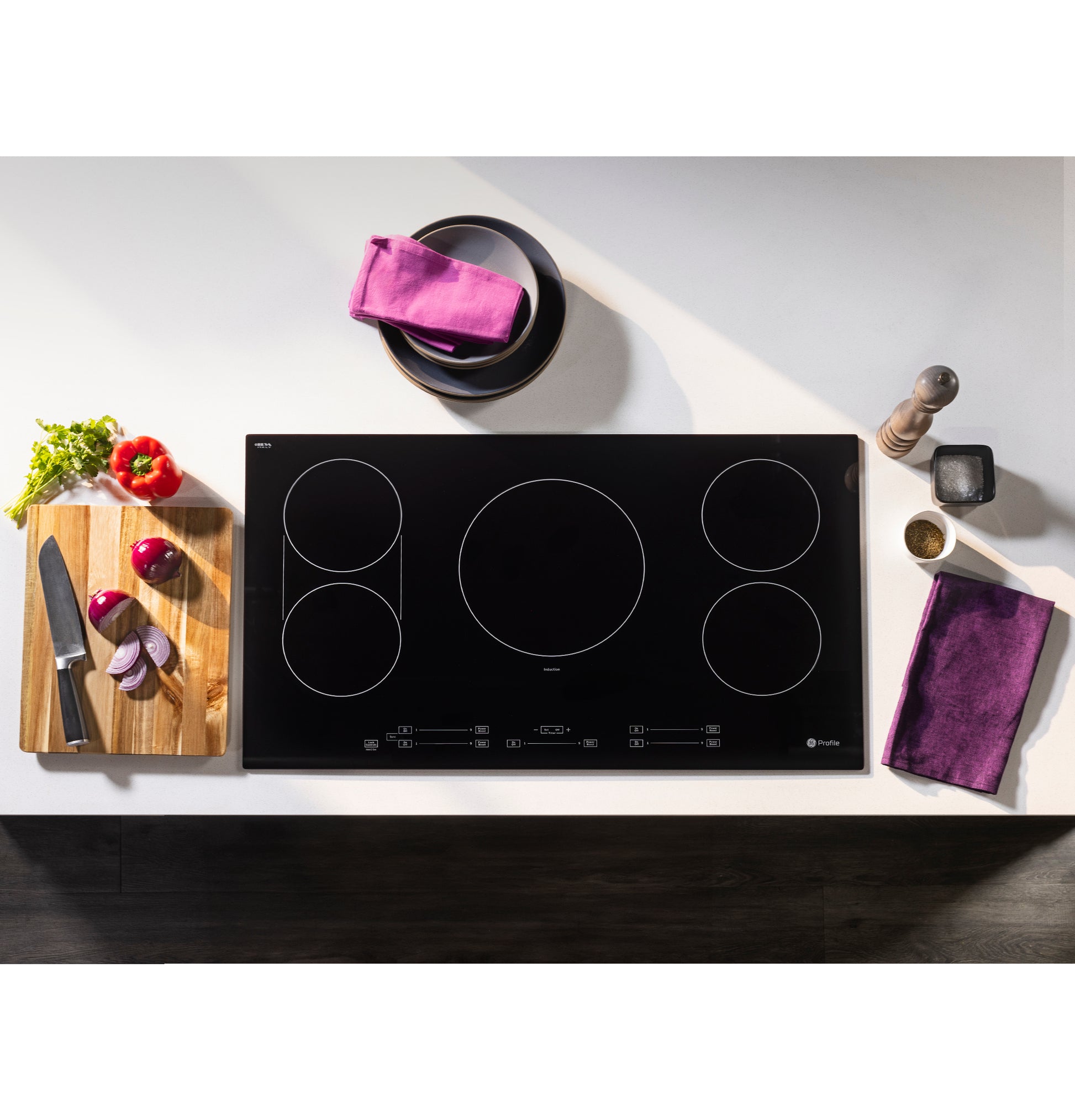 Sleek black touch control induction cooktop with five cooking zones on white countertop with fresh vegetables and kitchen tools