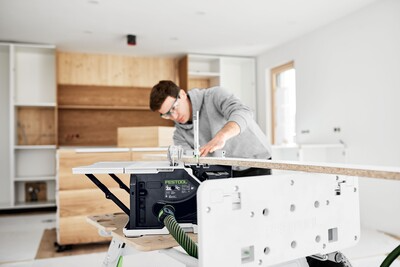 Man using a Festool cordless table saw to cut a wood board in a bright home improvement workshop