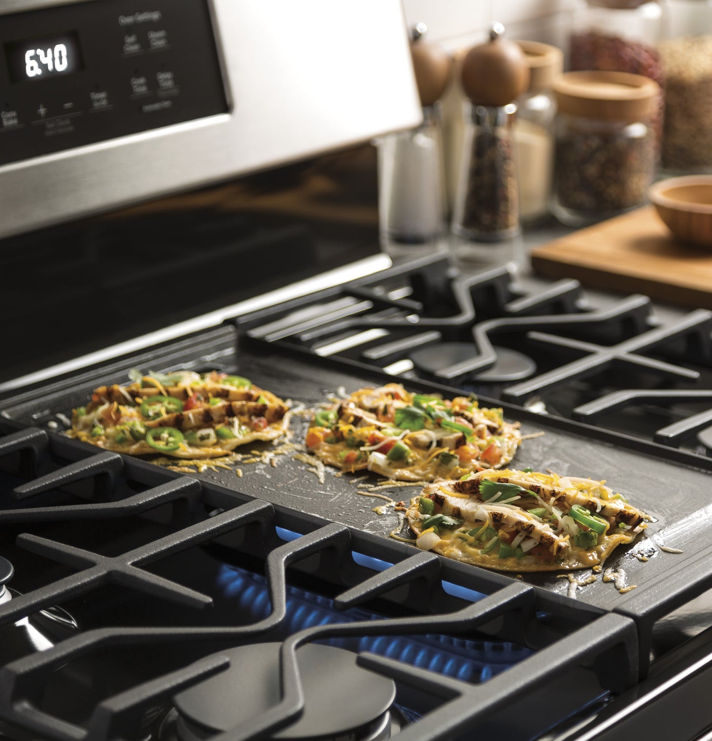 Gas burners on free-standing range with blue flames cooking three mini pizzas on griddle in modern kitchen setting