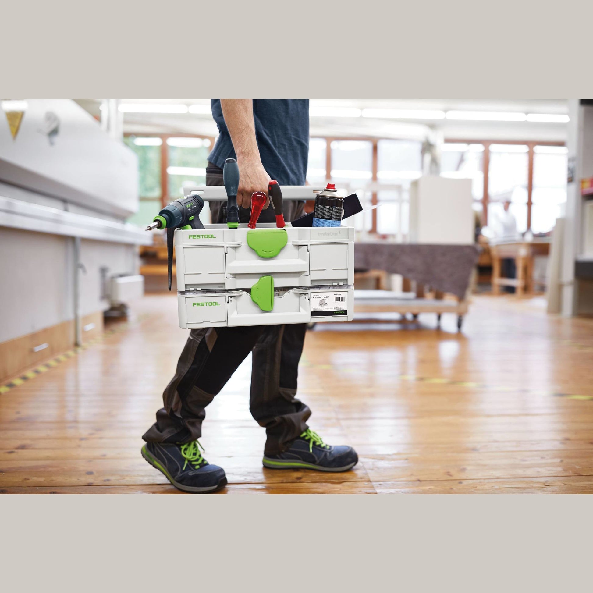 Person carrying white and green Festool modular toolbox with tools inside in a bright workshop setting