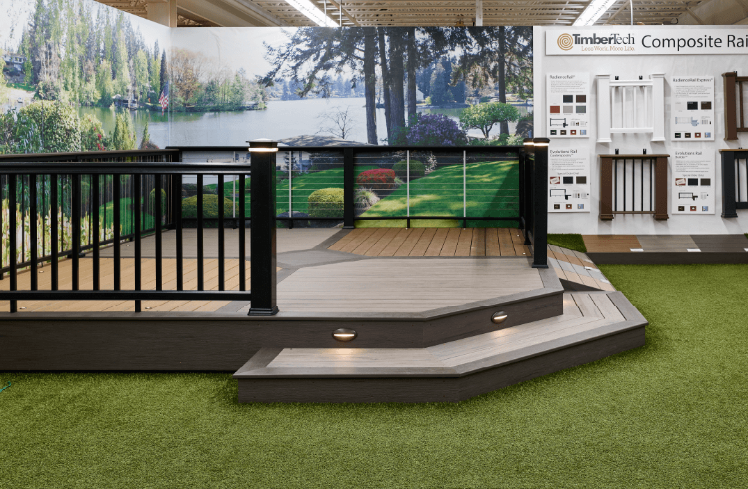 Indoor showroom display featuring a composite decking platform with black metal railing, tiered steps with built-in lights, and a backdrop of a lakeside forest scene.