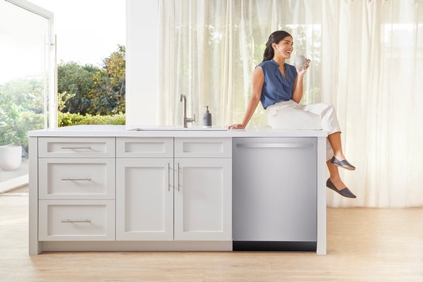 Woman sitting on modern kitchen island next to a stainless steel dishwasher with natural light and white cabinetry