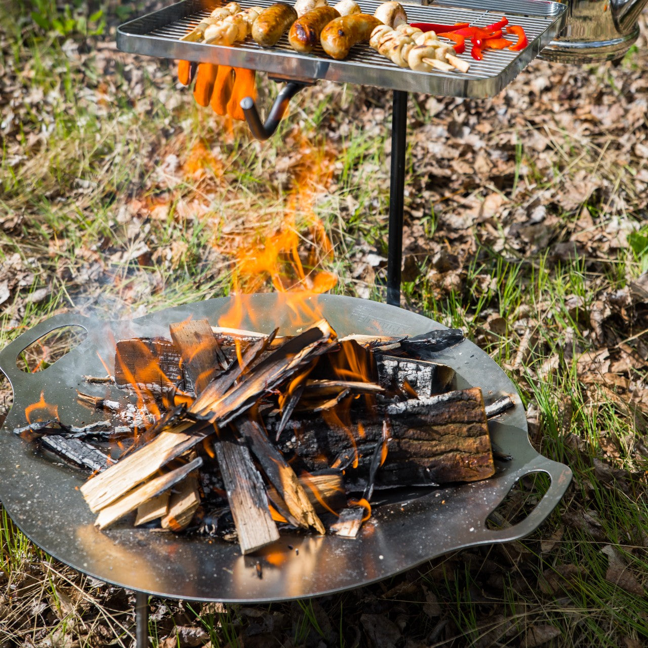 Outdoor fire bowl with burning wood and grilling rack cooking sausages and vegetables in grassy campsite setting
