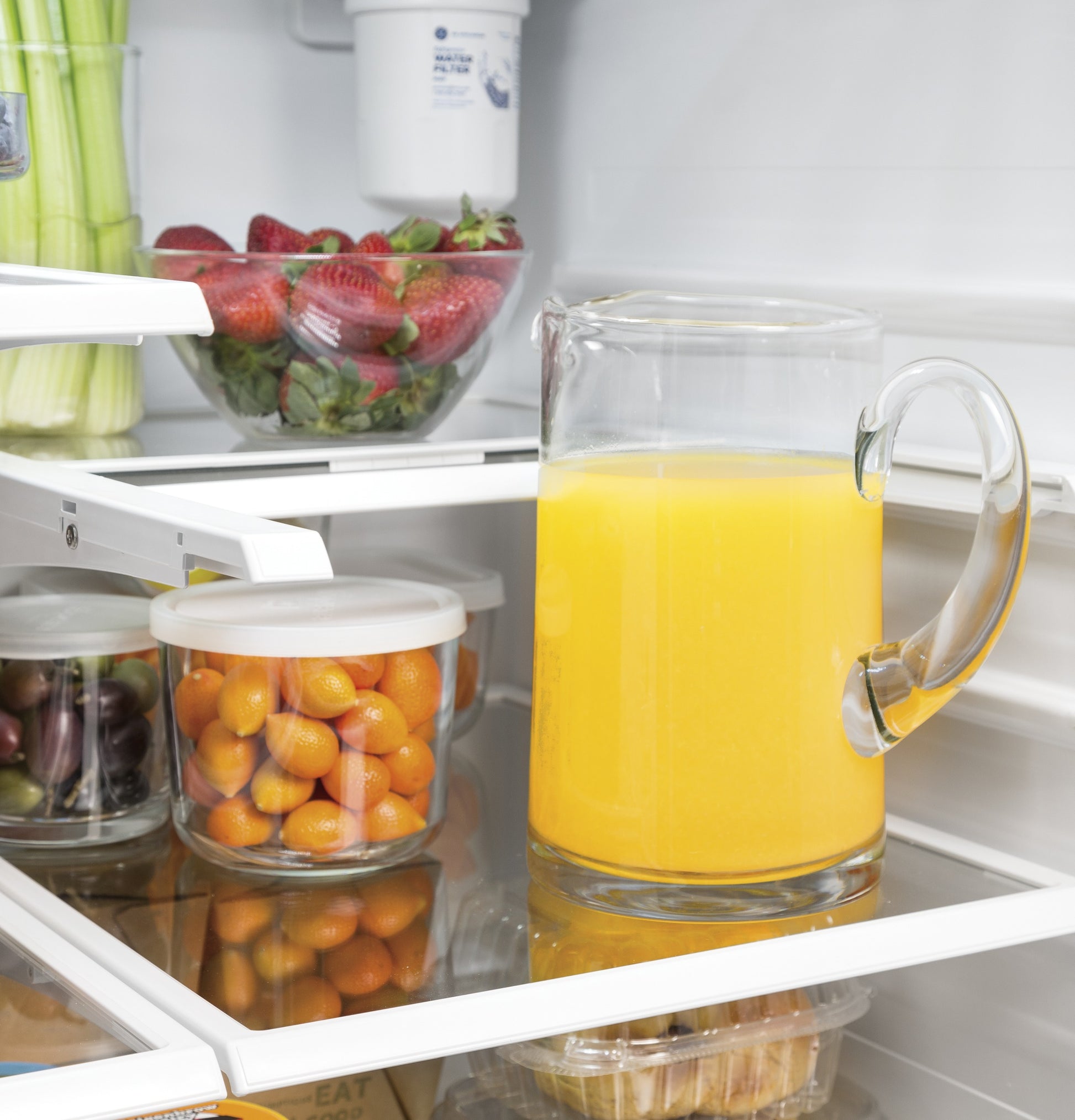Glass pitcher of orange juice and bowls of fresh fruit on glass shelves inside refrigerator in Appliances collection