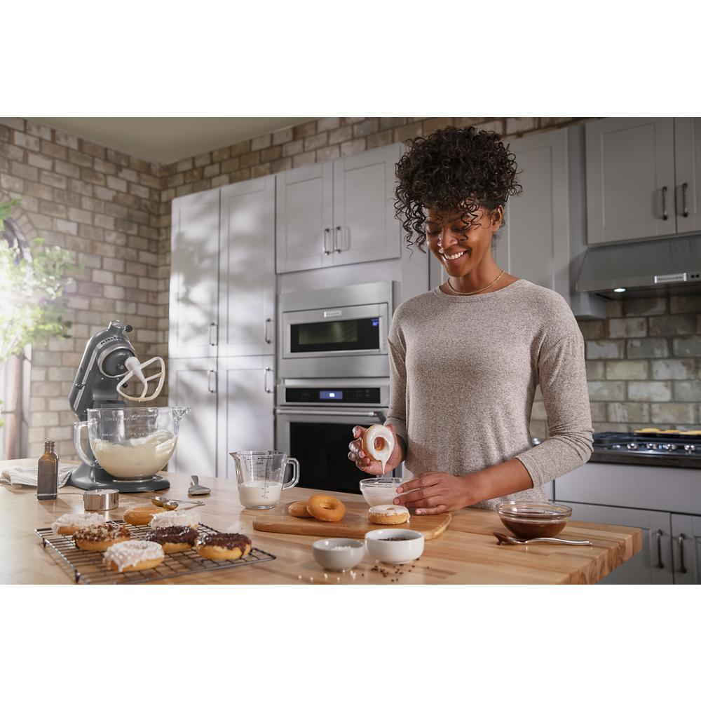 Bright kitchen with gray cabinets, stainless steel appliances, brick backsplash, and a woman decorating donuts on wooden island