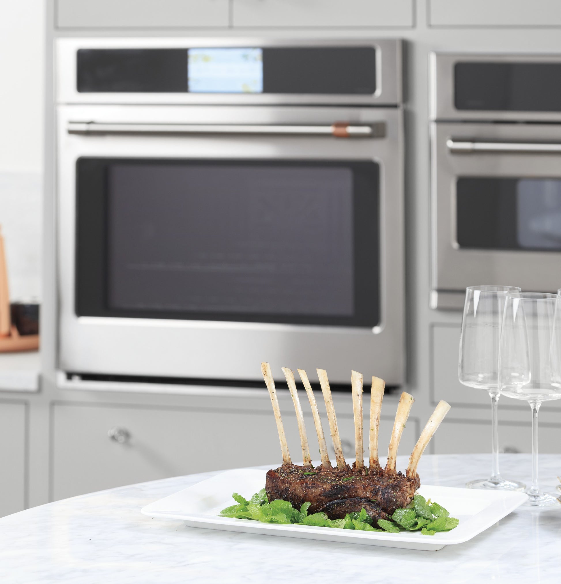 Rack of cooked lamb chops plated on herbs in front of stainless steel double wall oven in modern kitchen setting