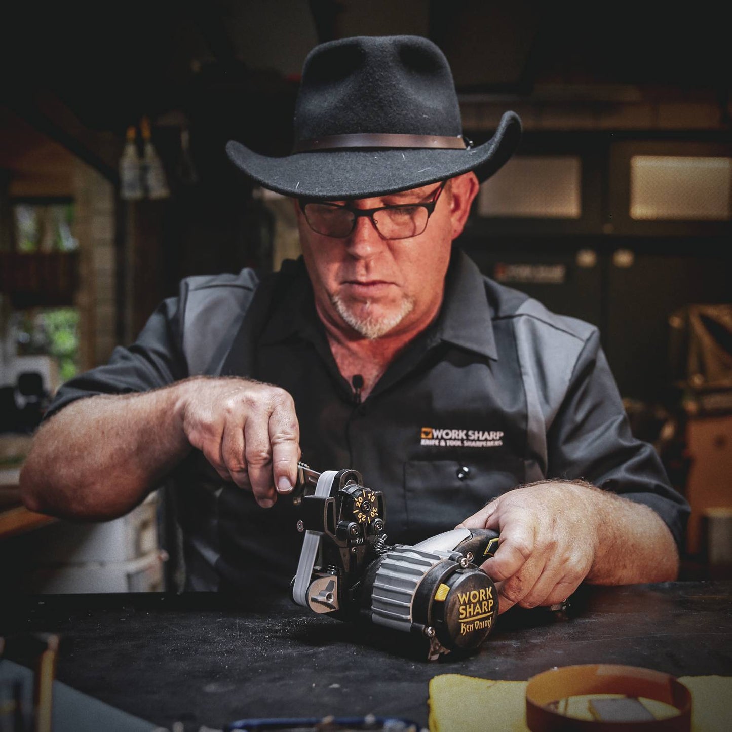 Man wearing black hat and glasses holding a Work Sharp Ken Onion tool sharpener on a workshop table