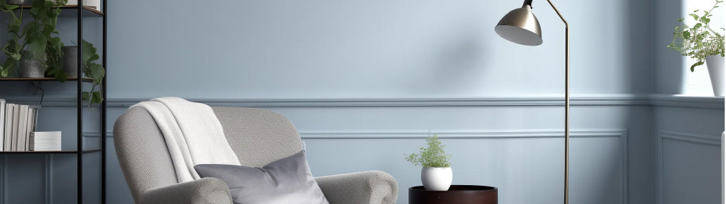 Cozy reading nook with a cushioned armchair draped with a throw, a small side table holding a plant, a tall floor lamp, and soft blue-gray walls with decorative molding.