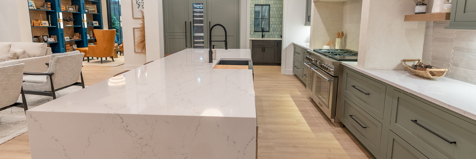 Spacious modern kitchen featuring a large white marble island, built-in stainless appliances, gray cabinetry, and light wood flooring with soft ambient lighting.