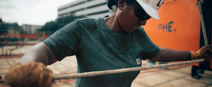 Worker wearing protective gloves, sunglasses, and a hard hat gripping a rope on an outdoor construction site with equipment in the background.