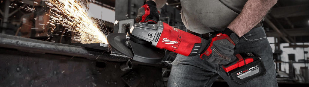 Worker wearing gloves using a cordless angle grinder to cut metal, creating bright sparks in an industrial workshop setting.
