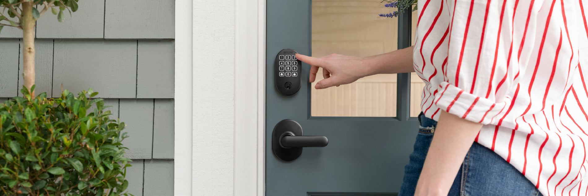 Person pressing the keypad on a dark-colored smart door lock installed on a blue exterior door with white trim and nearby green shrubbery.