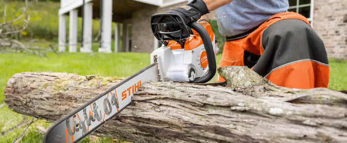 Person wearing protective gear using an orange and white chainsaw to cut a large log outdoors in a backyard setting near a brick house.