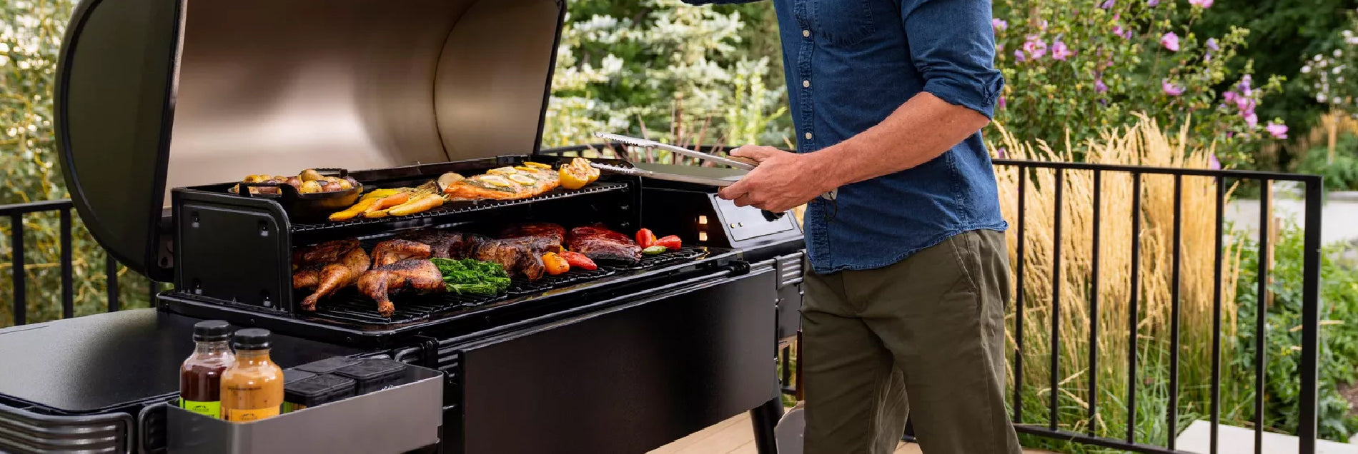 Person grilling an assortment of meats and vegetables on a large outdoor smoker grill with a raised lid, set on a patio surrounded by greenery.