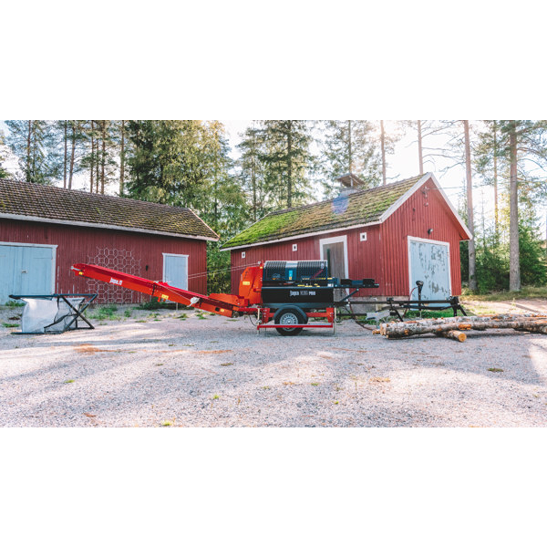 Red and black Japa 365+ ROAD firewood processor with conveyor belt near red wooden buildings in outdoor setting