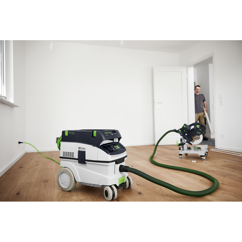 Compact dust extractor equipped with green hose and protective wheel covers on hardwood floor in workshop setting