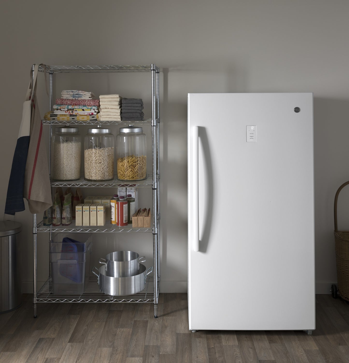 White frost-free upright freezer with vertical handle next to metal shelving stocked with kitchen supplies and cookware