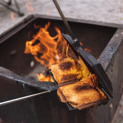 Double cast iron sandwich cooker with wooden handle toasting sandwiches over an open campfire for outdoor meal prep