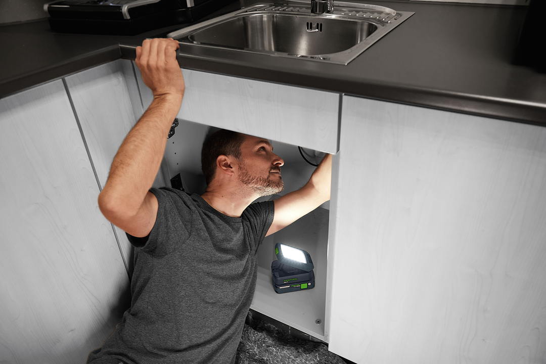 Man using compact Festool cordless light to illuminate plumbing area under sink for home improvement tools