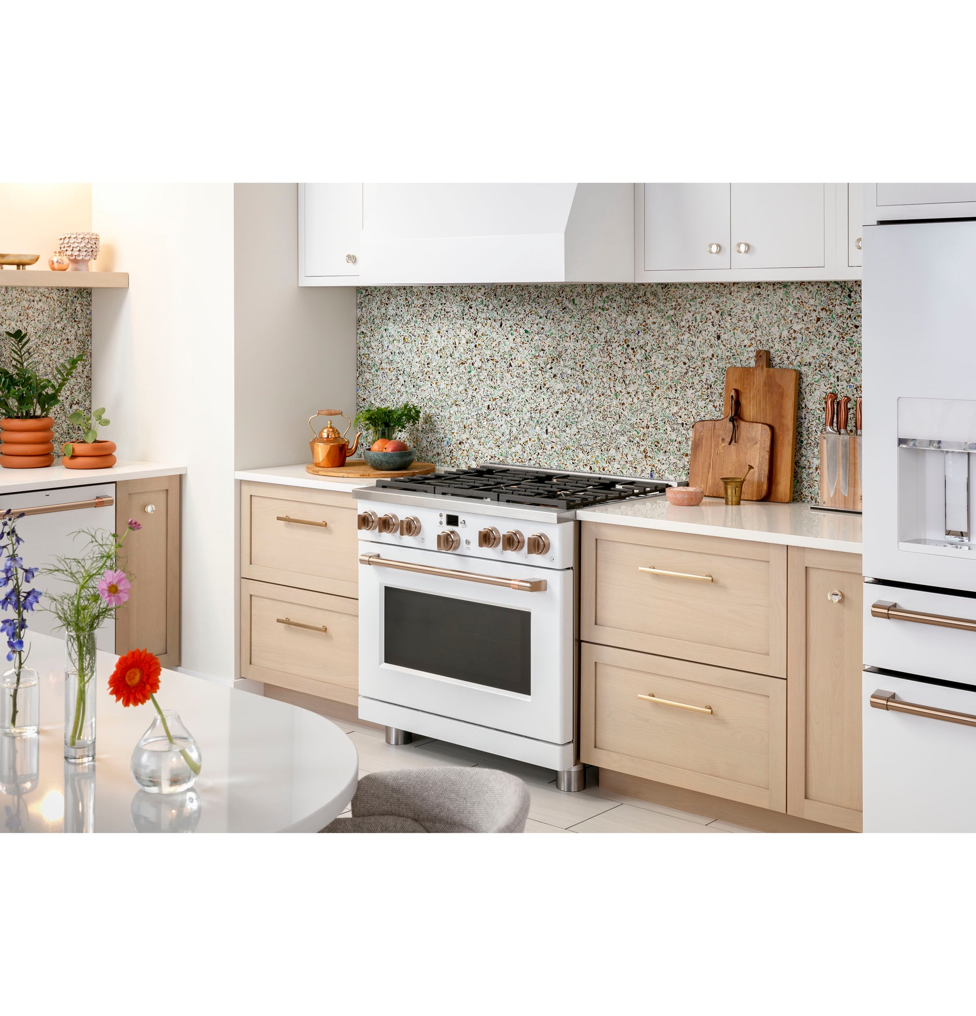 White all-gas kitchen range with six brass knobs set in light wood cabinetry and speckled stone backsplash