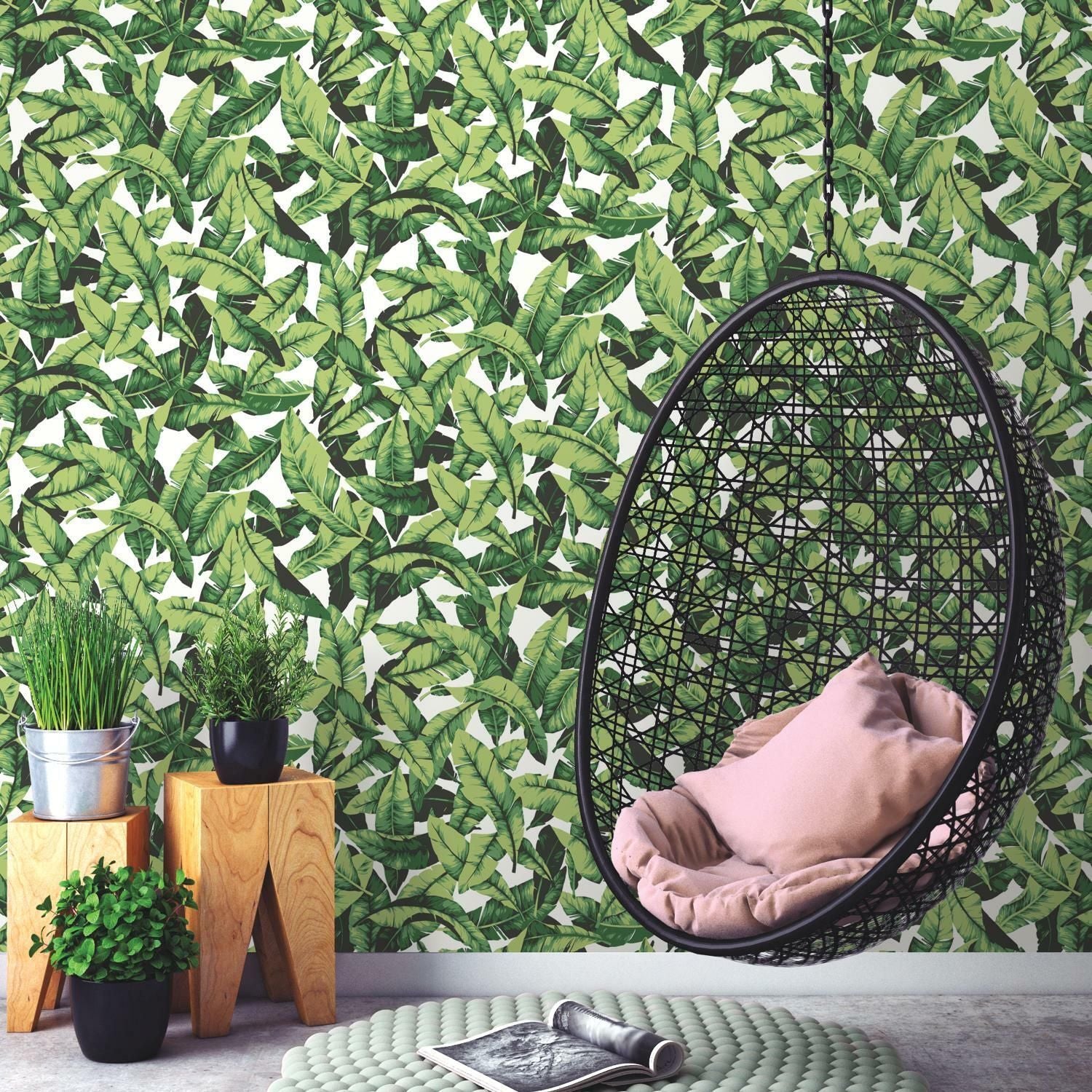 Indoor hanging black rattan chair with cushions against green palm leaf patterned peel and stick wallpaper backdrop