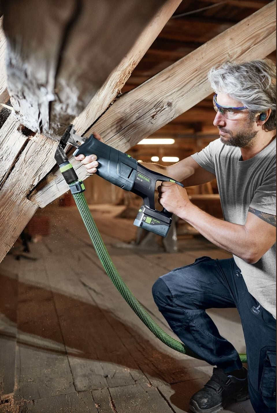 Man wearing protective glasses using cordless reciprocating saw to cut wooden beam indoors in home improvement setting