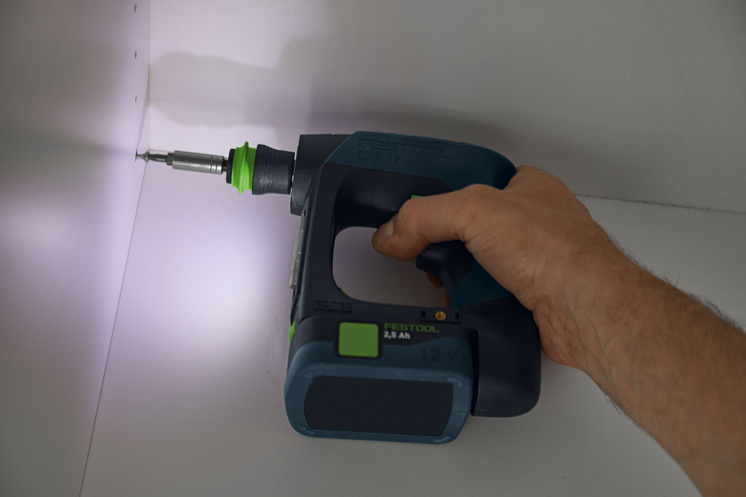 Hand holding compact cordless drill with LED light drilling into white cabinet corner in woodworking setting