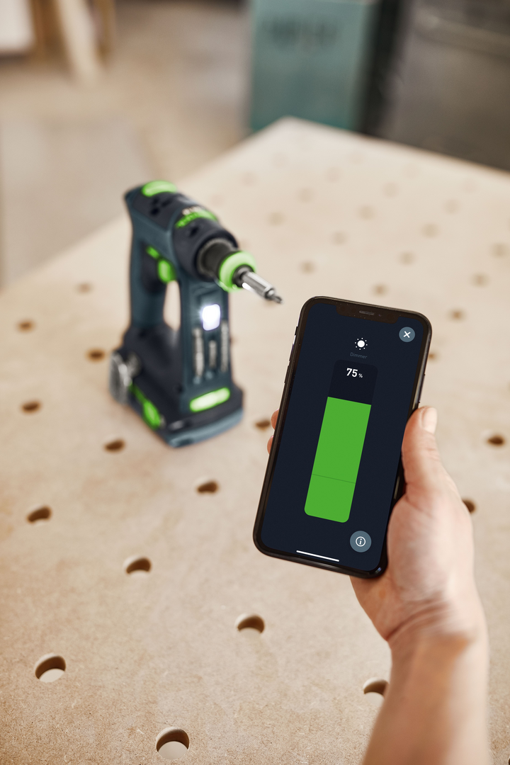 Hand holding smartphone with dimmer app controlling LED light on Festool cordless drill on workbench for woodworking tools