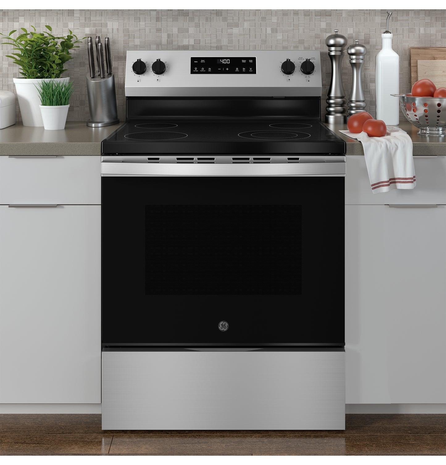 Stainless steel electric range with smooth black cooktop and control knobs in modern kitchen setting with accessories