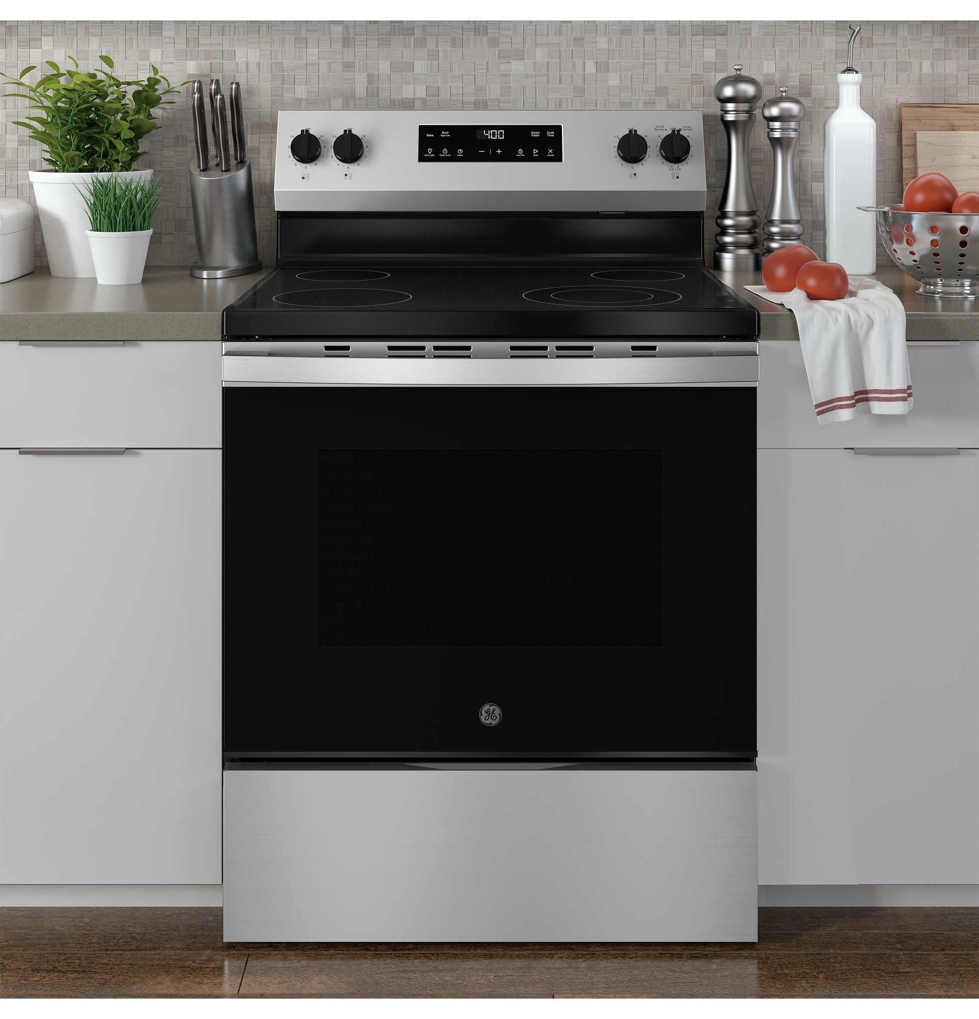 Stainless steel electric range with smooth black cooktop and control knobs in modern kitchen setting with accessories