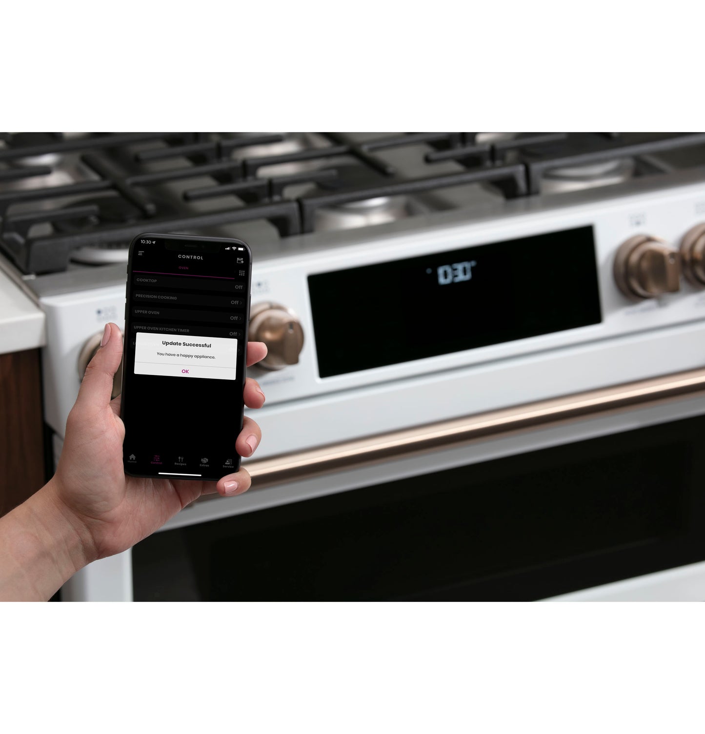 Hand holding smartphone showing update success message in front of white smart gas double-oven range with bronze knobs