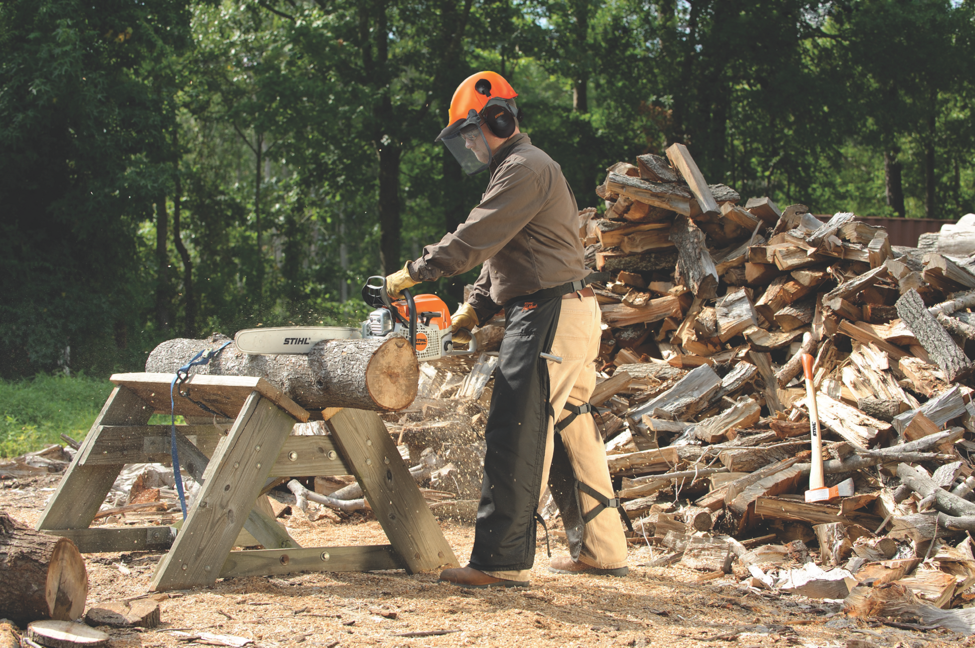 Person wearing safety gear using a gas-powered chainsaw to cut a log near a large pile of split firewood outdoors