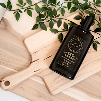 Black bottle of cutting board oil on light wood boards with green leafy branch in cleaning supplies collection under $50
