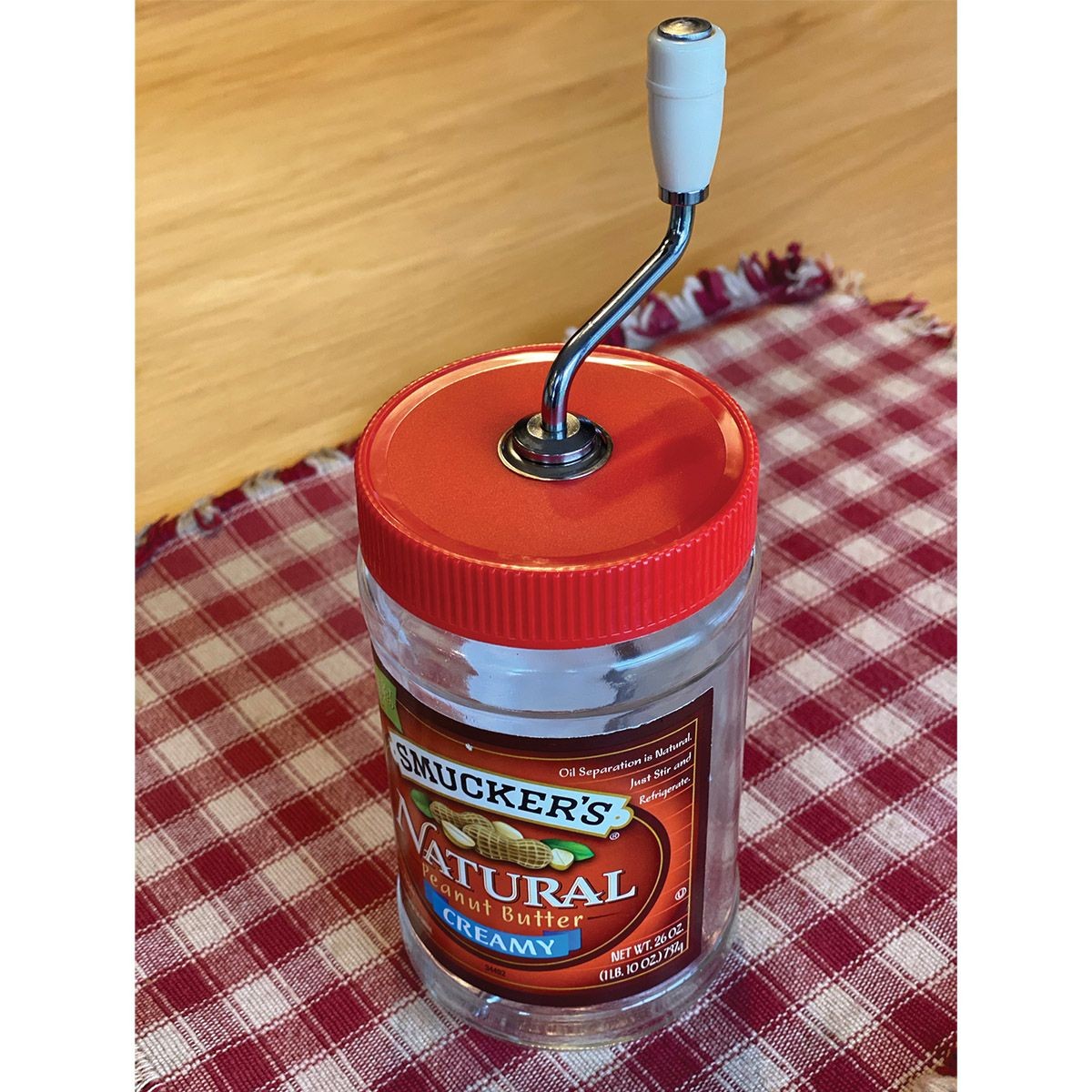 Red old fashioned peanut butter stirrer with metal crank handle and white grip atop jar of creamy peanut butter on red and white checkered cloth, in Kitchenware and Cooking Utensils under $50 collections.