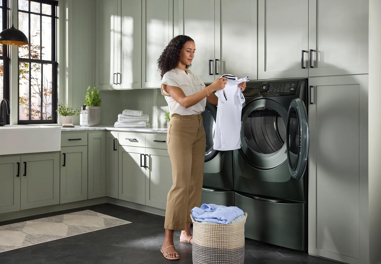 Woman holding a white shirt near a dark green front load electric dryer in a bright modern laundry room with green cabinetry