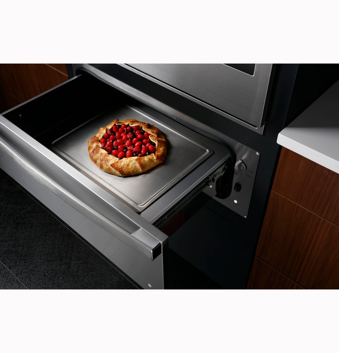 Open stainless steel warming drawer in modern kitchen with berry galette on baking tray inside appliance drawer
