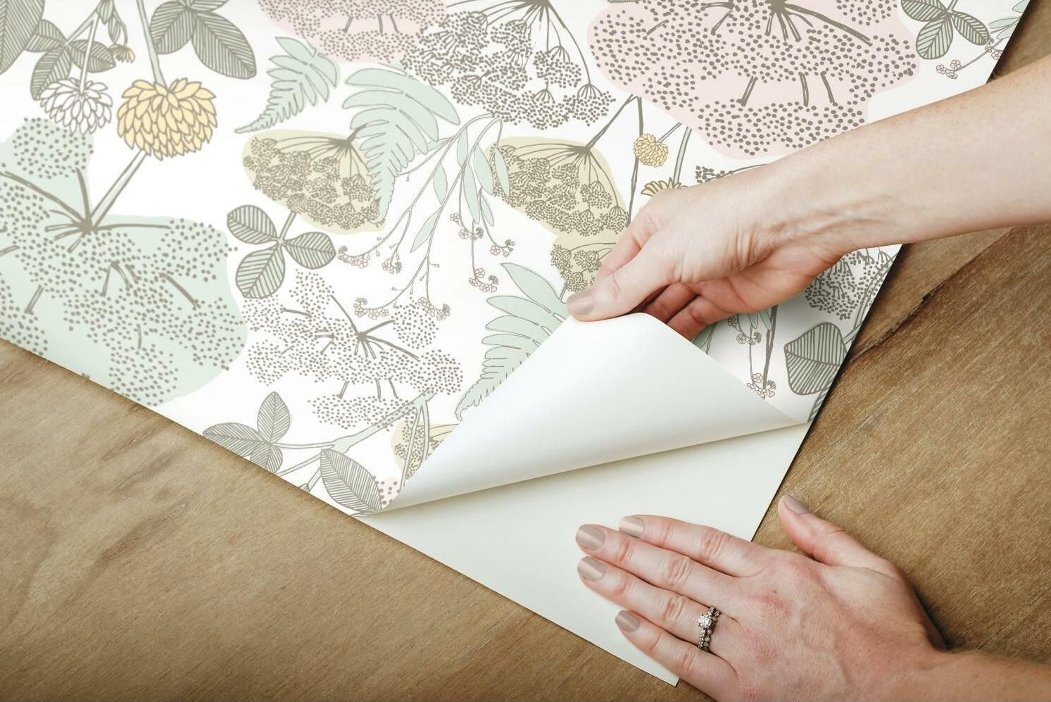 Hands peeling back floral patterned pink and green removable wallpaper on wooden surface for home decor