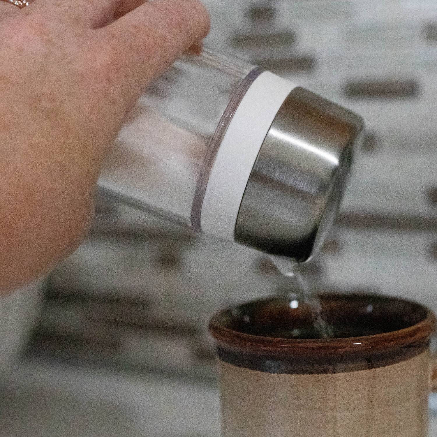 Hand pouring sugar from a clear dispenser with stainless steel lid into a brown ceramic cup in kitchenware setting