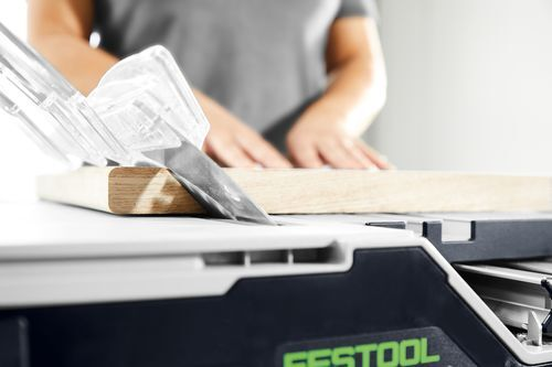 Person using a cordless Festool table saw with safety guard to make a precise cut on a wood board in a workshop