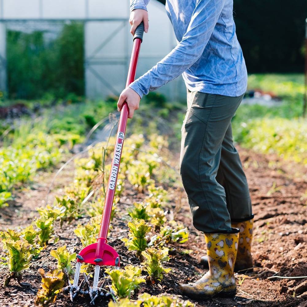 Person using a Garden Weasel steel cultivator with red handle tilling soil in outdoor garden under daylight