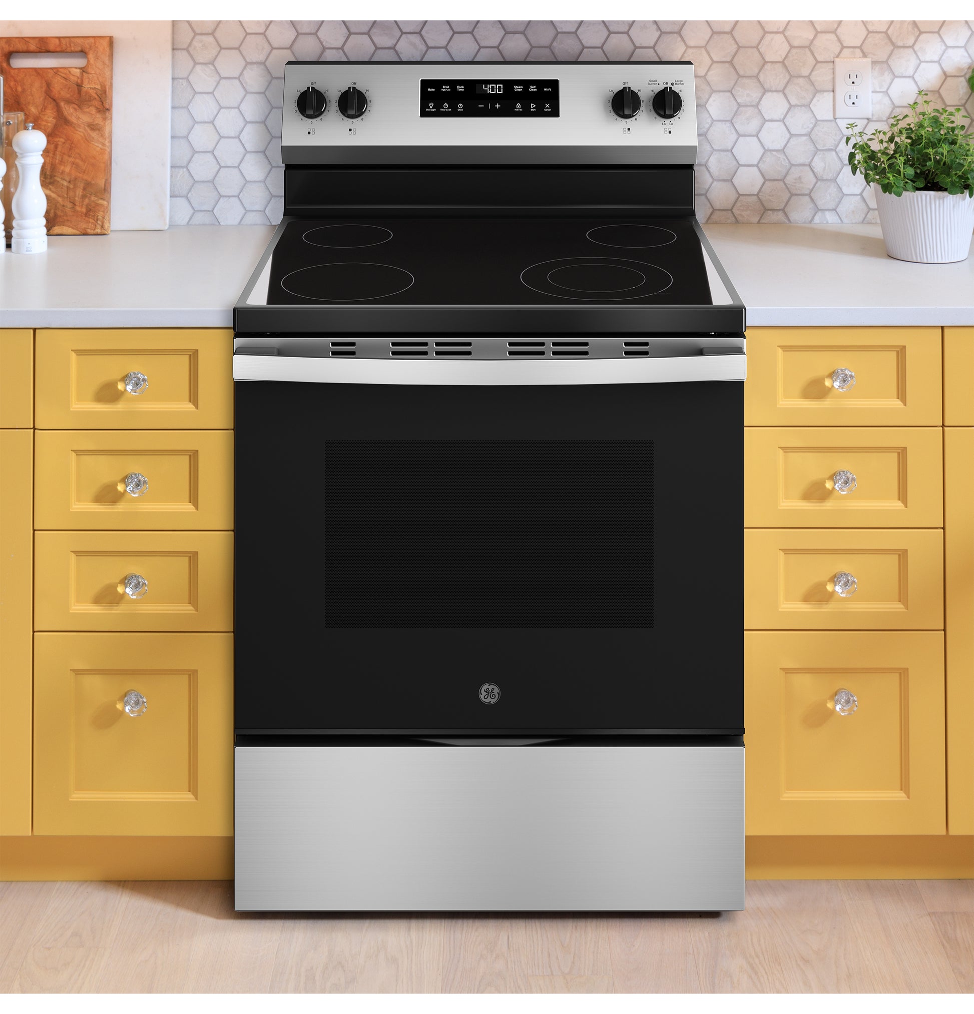 Stainless steel and black free-standing electric range set between yellow kitchen cabinets with hex tile backsplash and countertop accessories