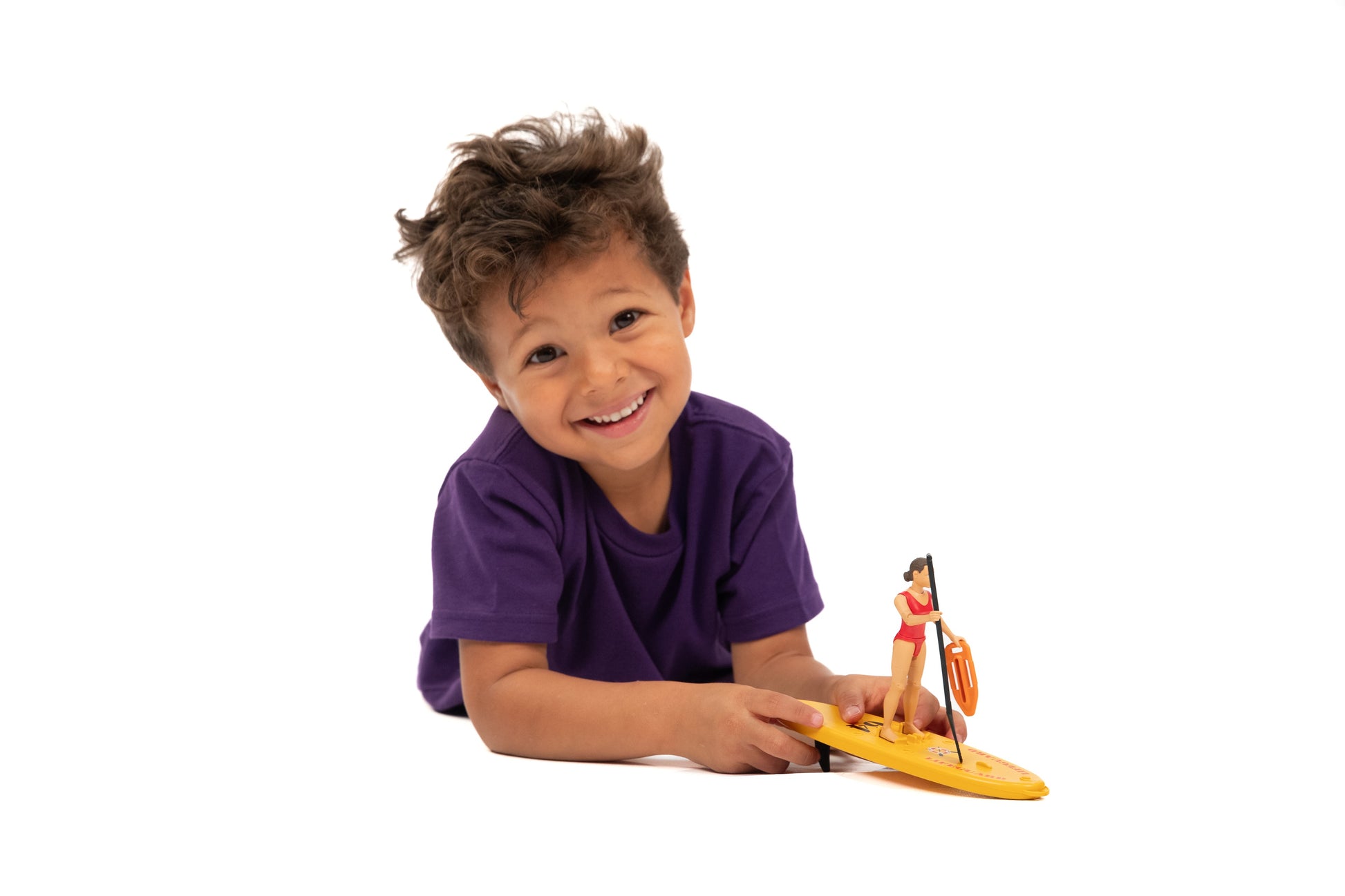 Smiling child playing with a lifeguard figure on a yellow stand-up paddleboard toy against white background