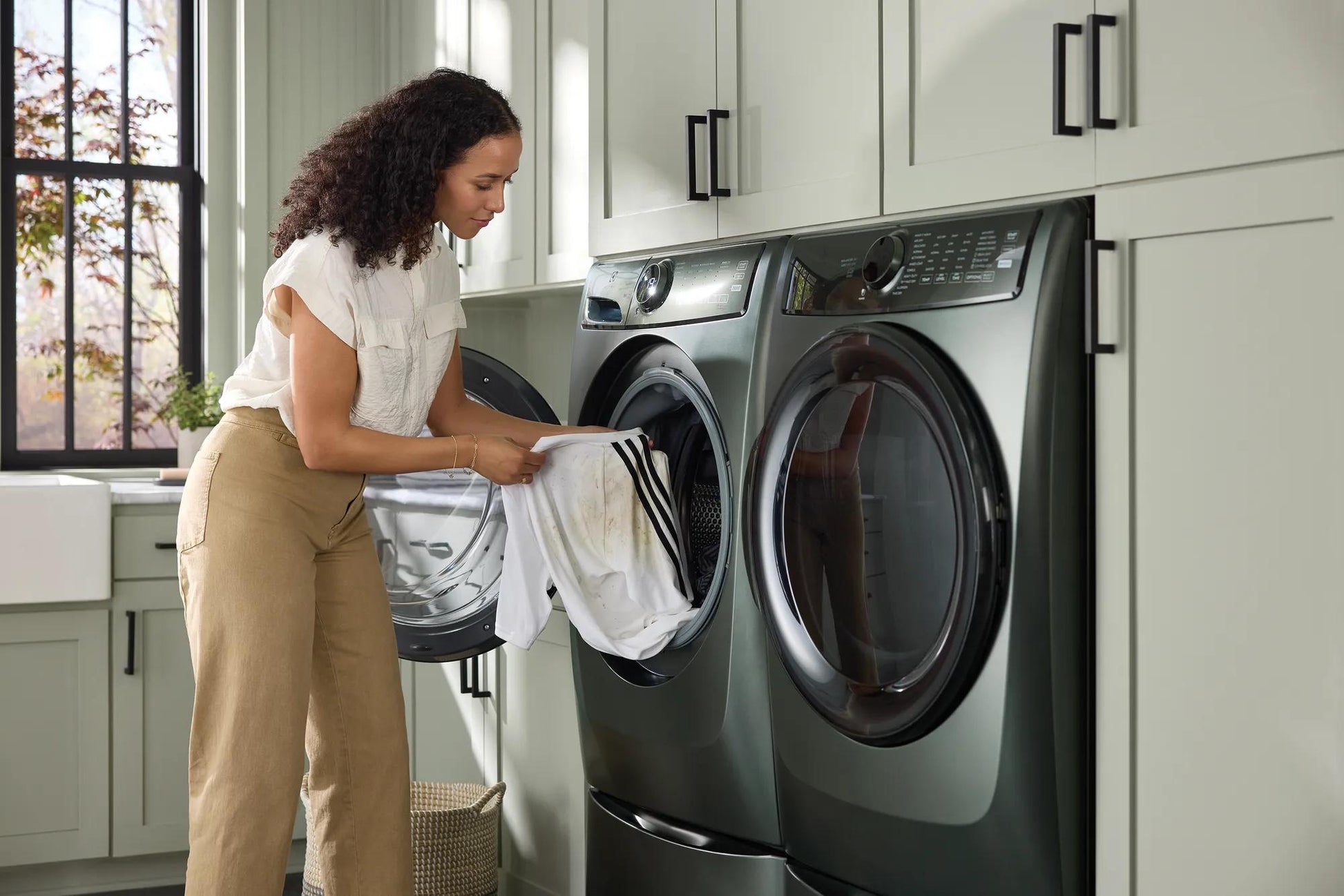 Woman loading laundry into a dark green front load electric dryer in a modern laundry room with gray cabinetry and natural light