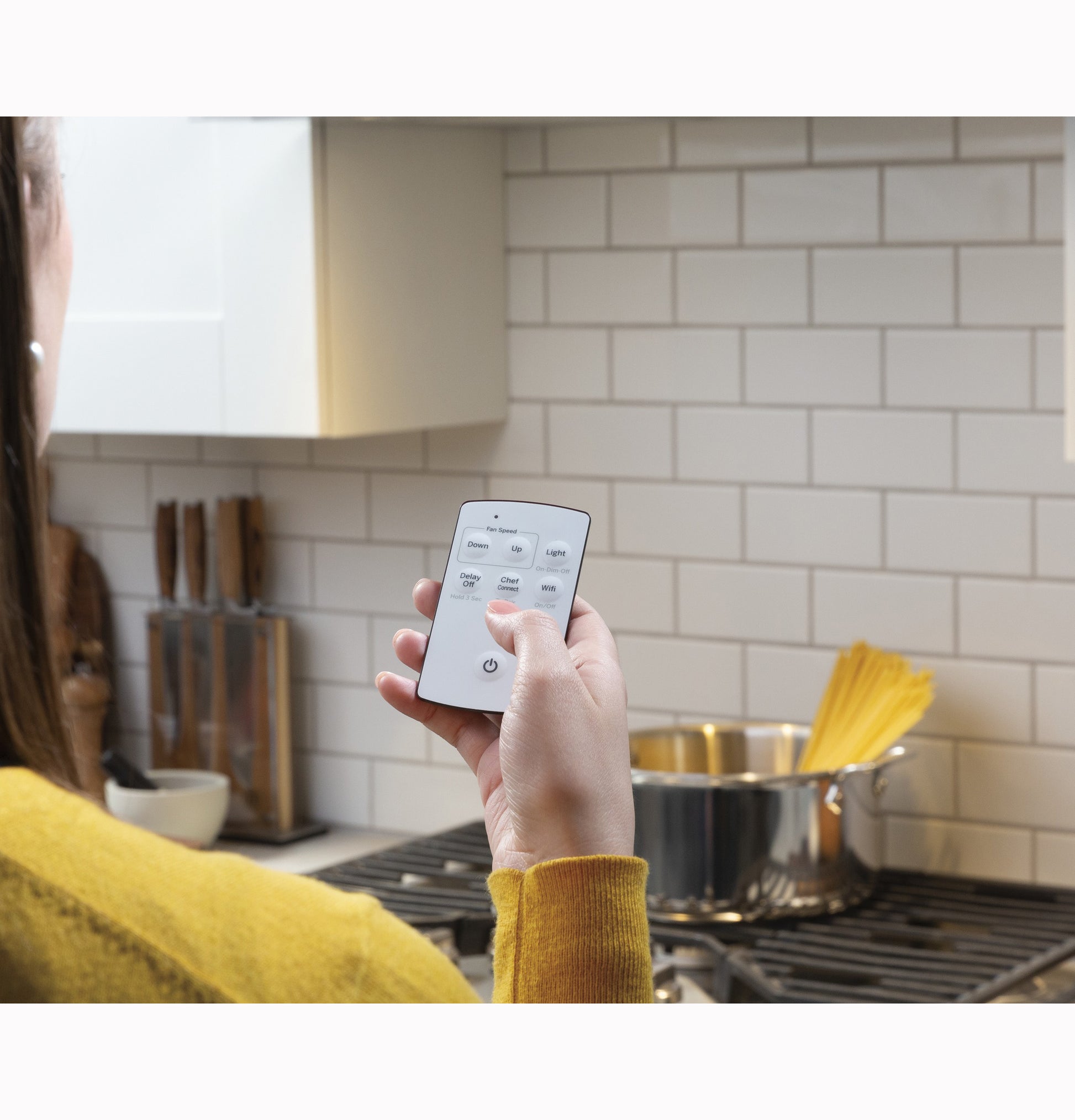 Person using white remote control to operate kitchen wall-mounted range hood above stainless steel stove with pot and spaghetti